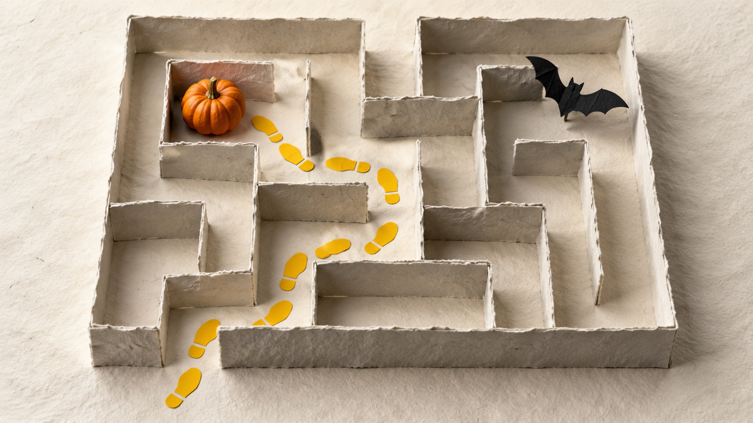 Folded paper maze, small pumpkin and black paper bat with a trail of bright yellow footprints.