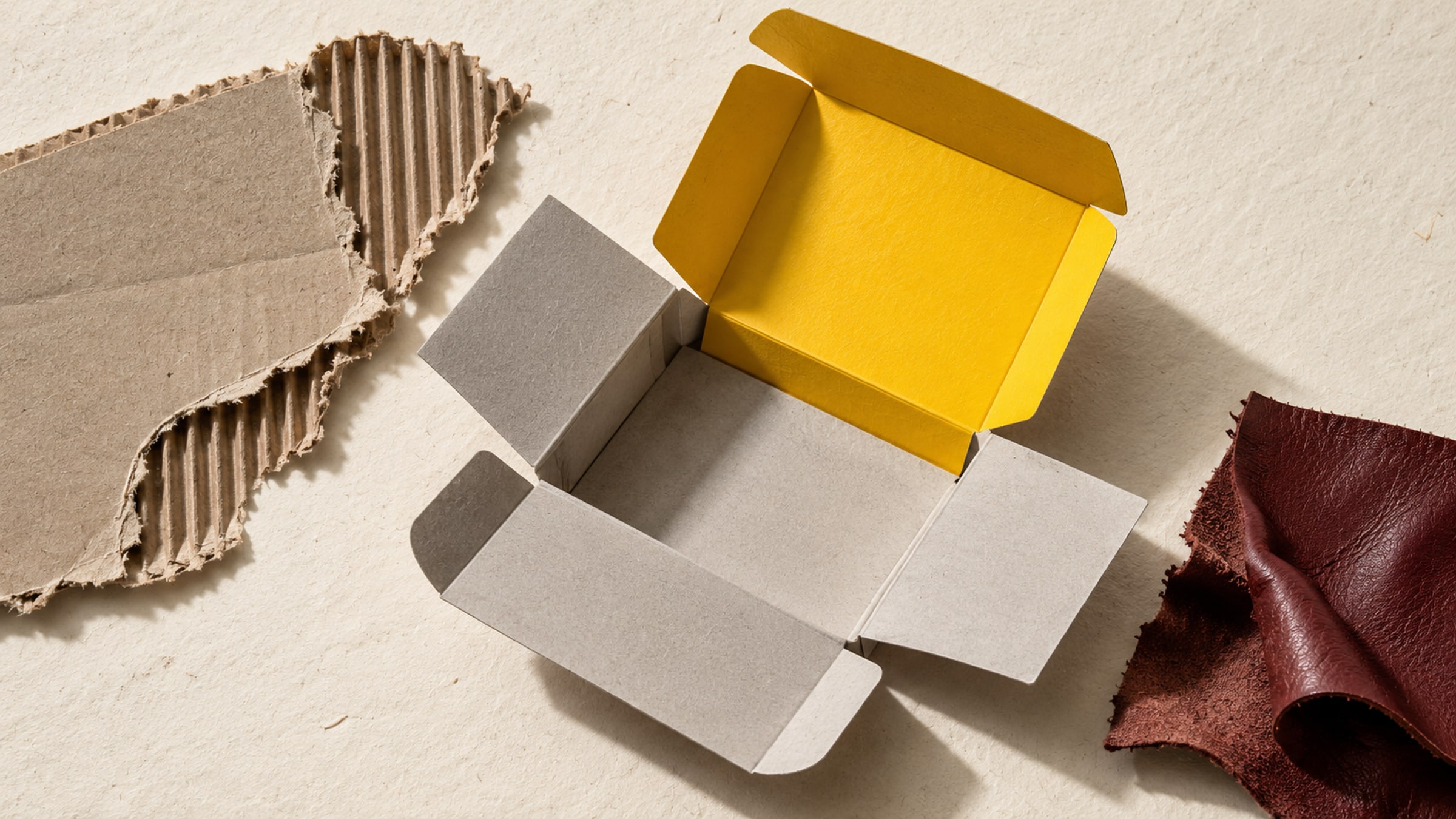 Unfolded monochrome cardboard carton with a yellow inner flap, corrugated paper and an oxblood leather offcut.